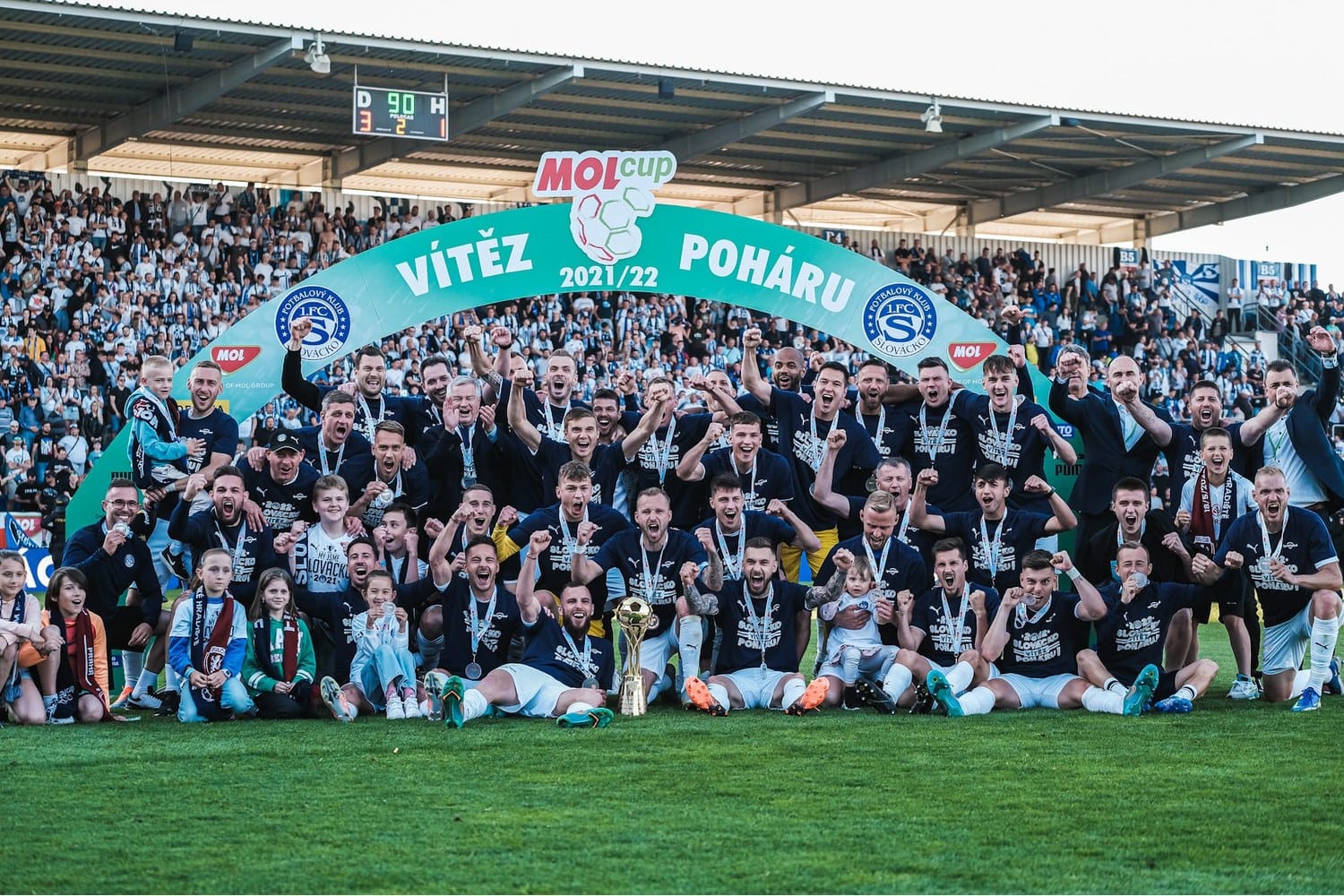 1.FC Slovácko players and staff pose for a team photograph with the 2021-22 Czech Cup trophy on the pitch under a green "VÍTĚZ POHÁRU 2021/22" arch, with the stadium stands full in the background.