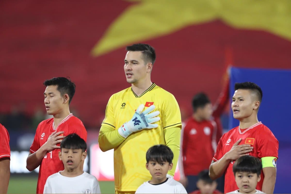 Filip Nguyen in a yellow Vietnam goalkeeper jersey standing for the national anthem with hand on heart, flanked by two teammates in red Vietnam shirts including captain Nguyen Quang Hai on the right, with a large Vietnamese flag in the background.