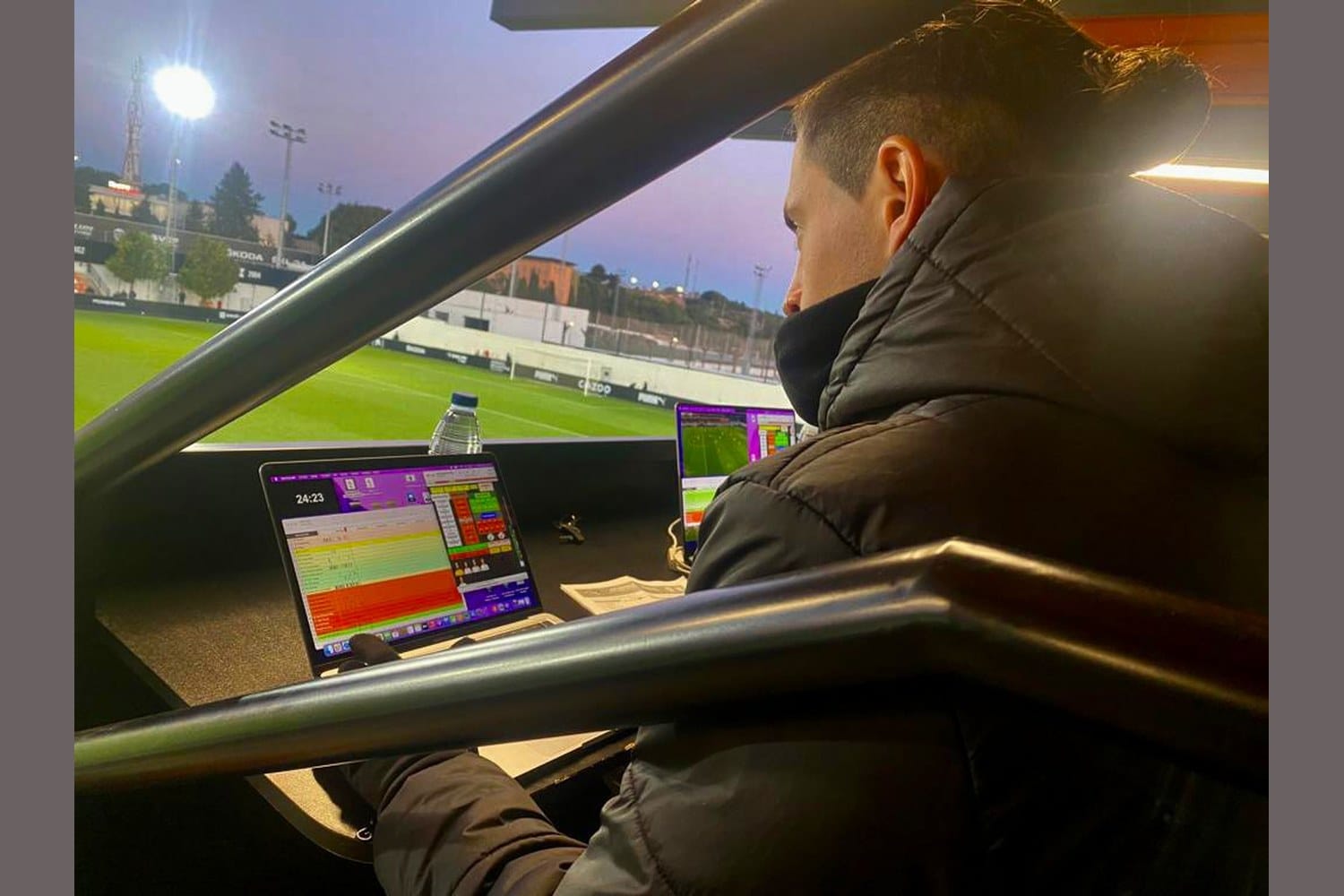 View from inside a stadium analyst box at dusk. A laptop in the foreground shows a HUDL Sportscode timeline in play; a second laptop beside it shows a live match feed. Nacho Alcaraz sits in profile, in a dark jacket, watching the pitch below.