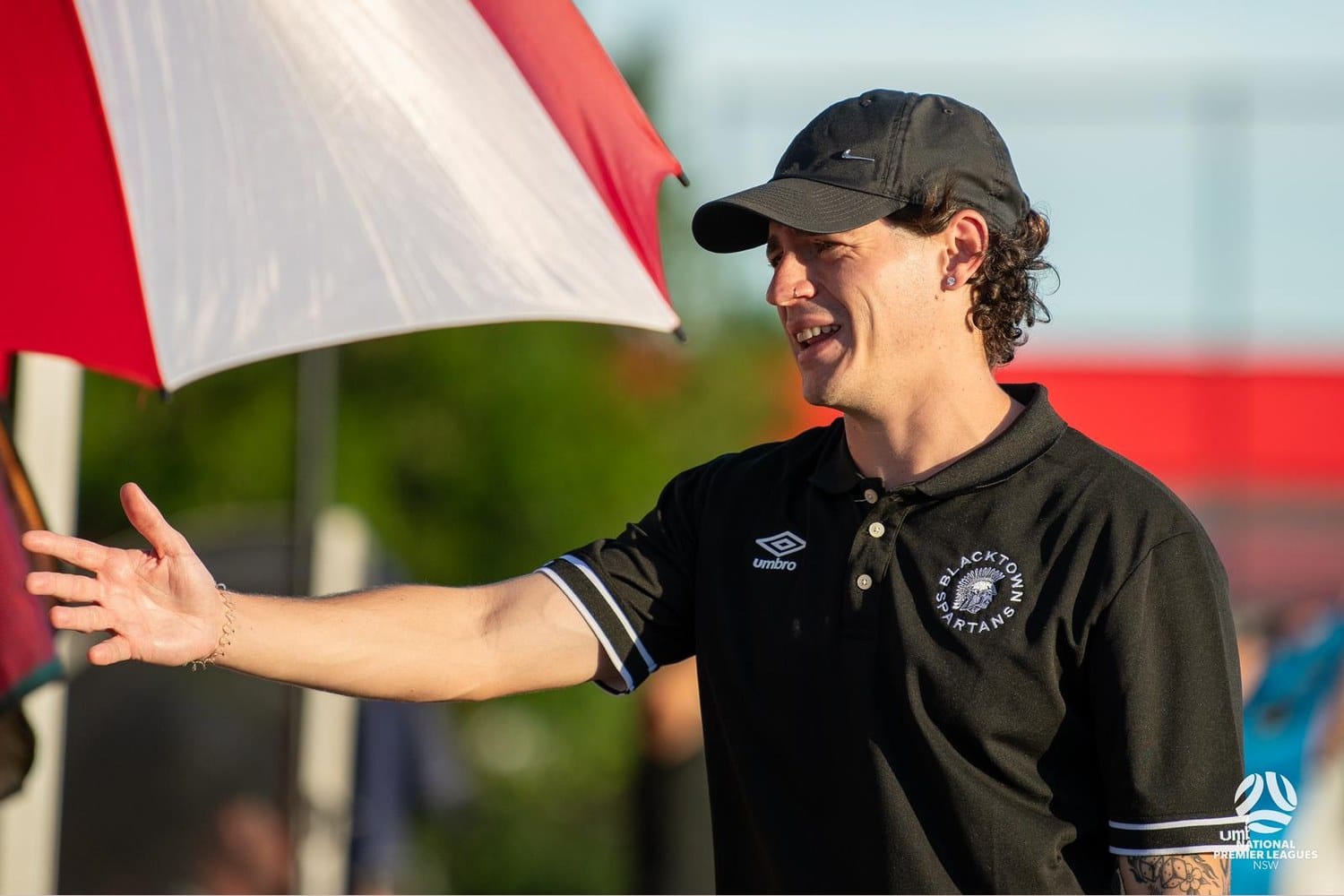 Nacho Alcaraz, in a black Blacktown Spartans polo shirt and black cap, stands on the sideline during an NPL NSW match in Australia, right arm extended as he gives on-pitch instruction.