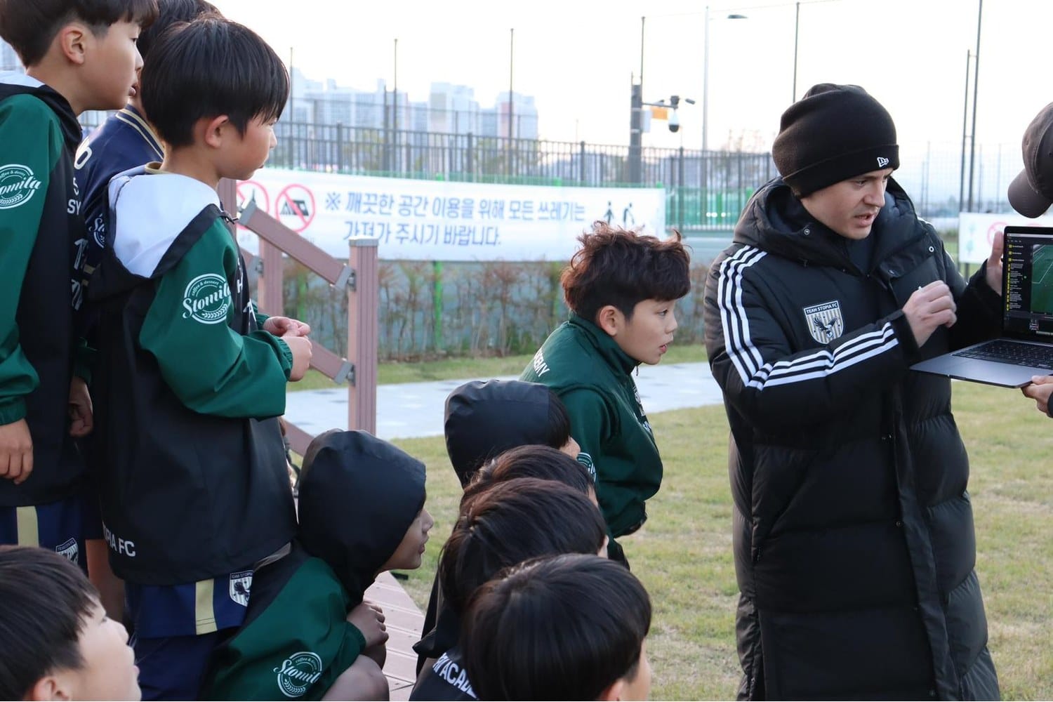 Nacho Alcaraz, in a black Team Stopia FC puffer jacket and beanie, stands at the touchline of a Korean youth training ground showing a laptop screen to a group of young Team Stopia FC players in green and navy kits.