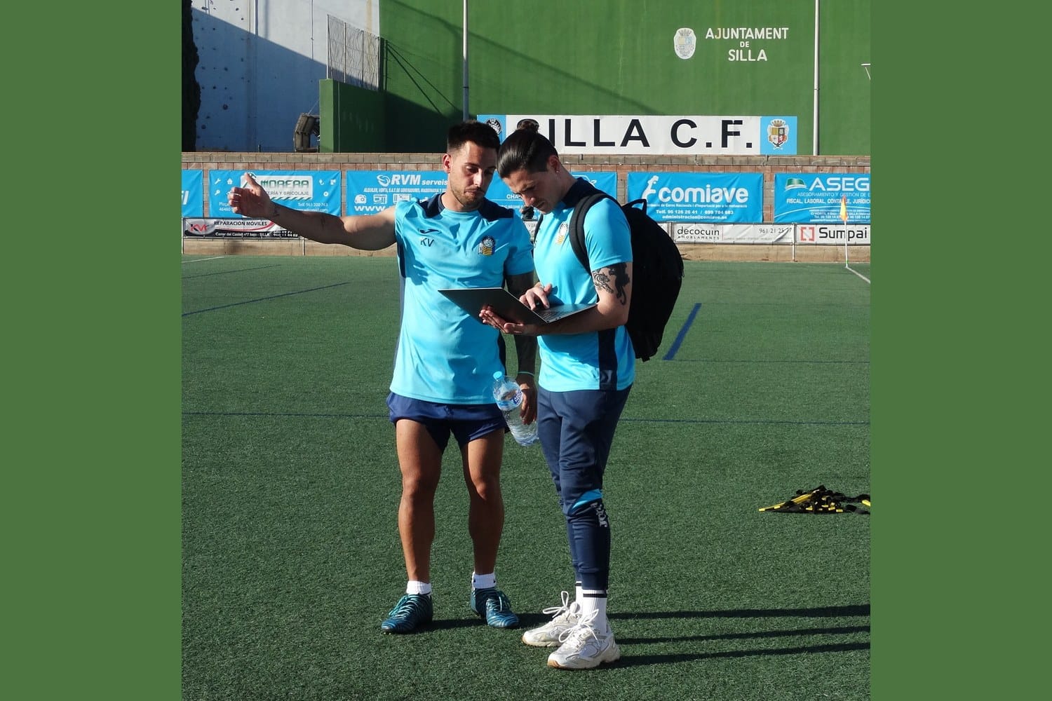 Nacho Alcaraz, in a Silla CF teal training kit with a backpack over his shoulder, reads from a laptop held open in his hands as a staff colleague beside him gestures with an outstretched arm, pointing out something on the training pitch.