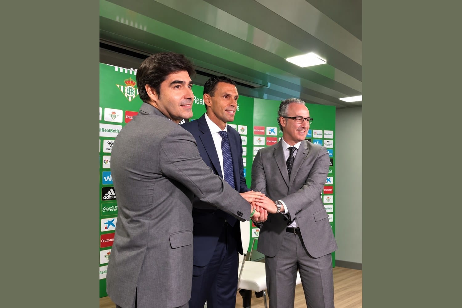 Miguel Torrecilla, on the right in a gray suit, with Real Betis president Ángel Haro at left and Gustavo Poyet in the centre, the three joining hands in front of a Real Betis sponsor backdrop.