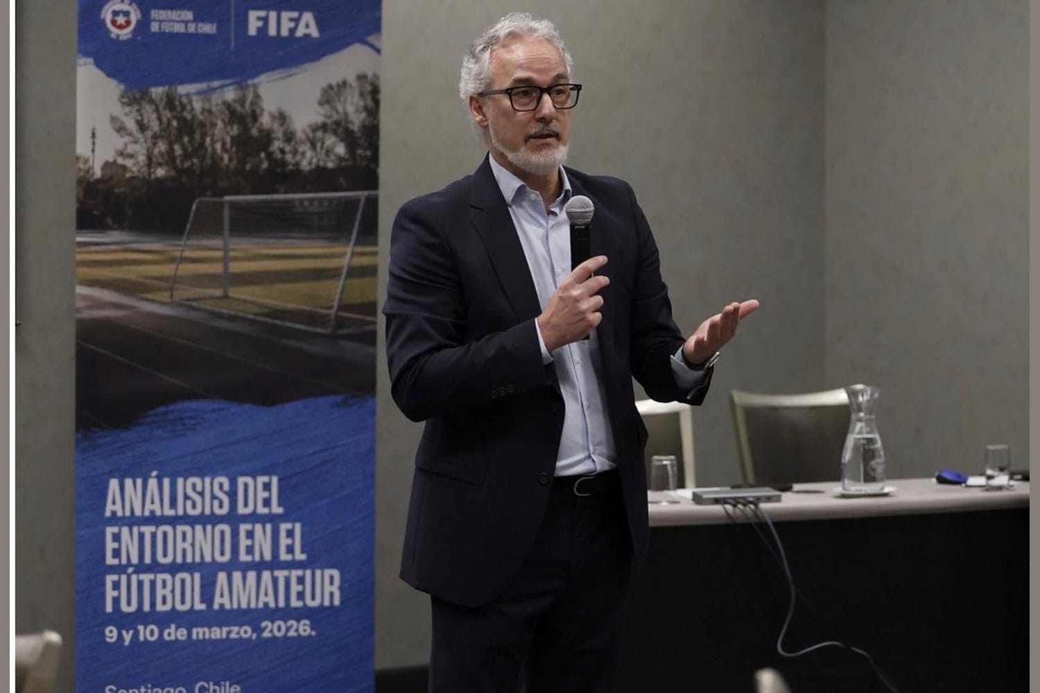 Miguel Torrecilla, standing in a dark suit, speaking with a microphone next to a Federación de Fútbol de Chile and FIFA banner reading "Análisis del Entorno en el Fútbol Amateur, 9 y 10 de marzo, 2026, Santiago, Chile."