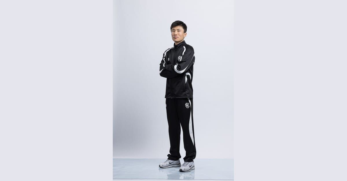 Ki-hyun Kim, Korean football coach and analyst, standing with arms crossed in a black tracksuit against a plain light-grey studio backdrop.