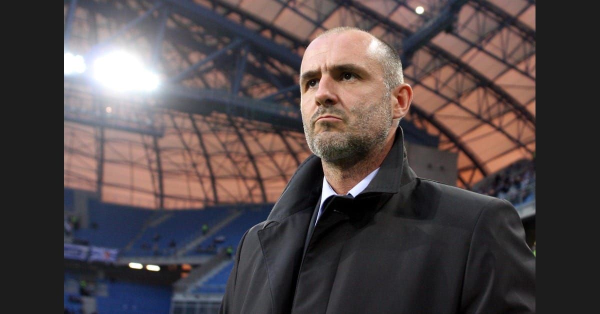 Michał Probierz, Polish head coach, in a dark overcoat on the touchline inside a stadium, looking upward with a composed, focused expression.