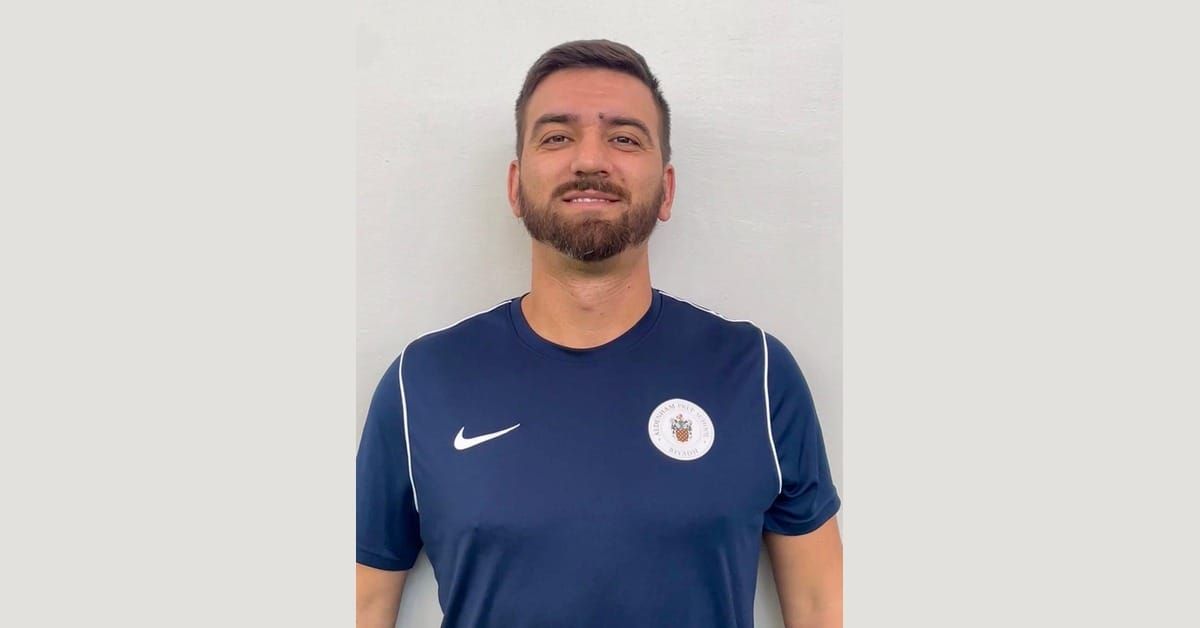 Simone Canepa, Italian football coach, in a navy Aldenham Prep School polo shirt, smiling in a head-and-shoulders portrait against a plain light wall.