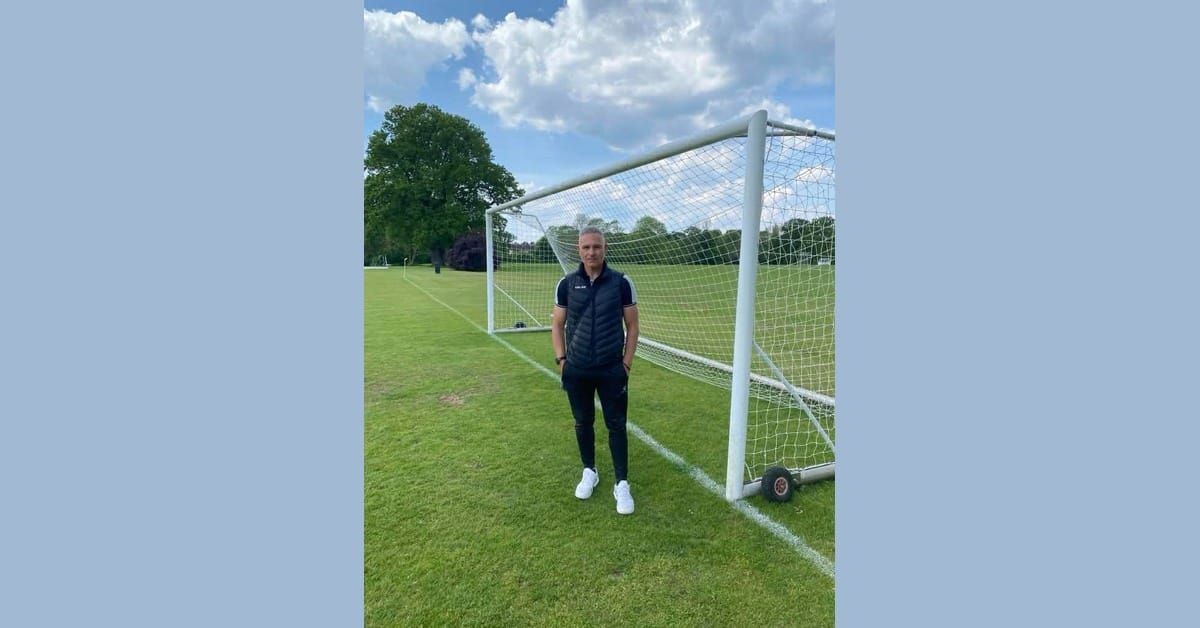 Lefteris Kalogirou, Greek football coach, standing in front of a goal on a grass pitch in a black gilet with hands in his pockets, under a partly cloudy sky.
