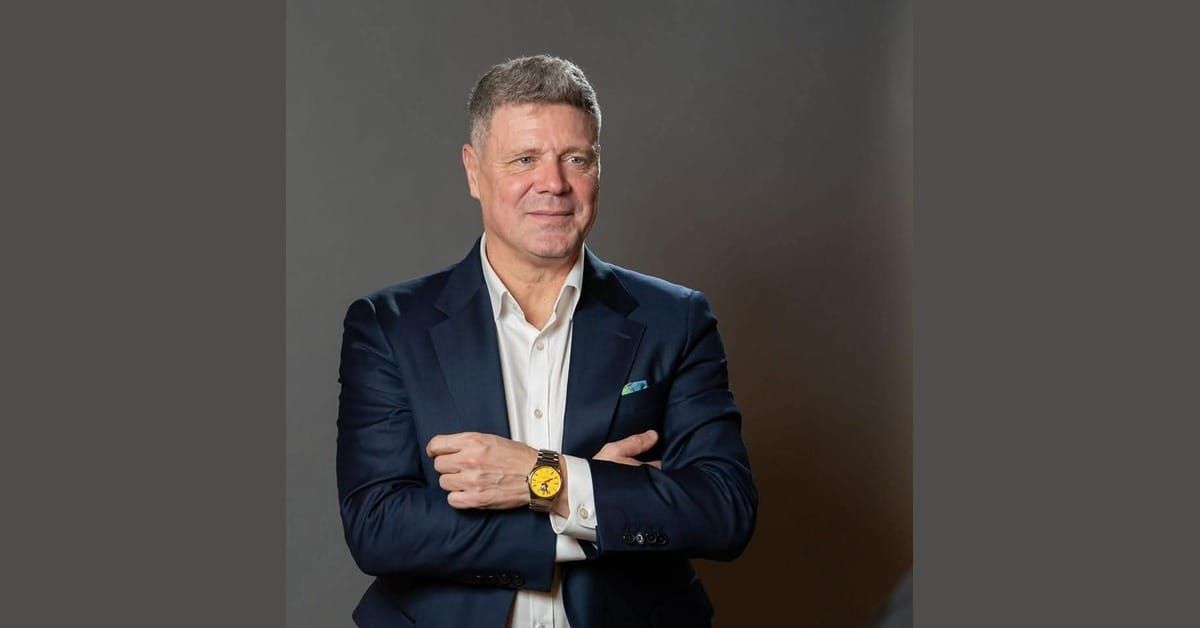 Tibor Selymes, Romanian football coach and former international, in a navy suit over a white shirt with arms crossed, in a studio portrait against a grey background.
