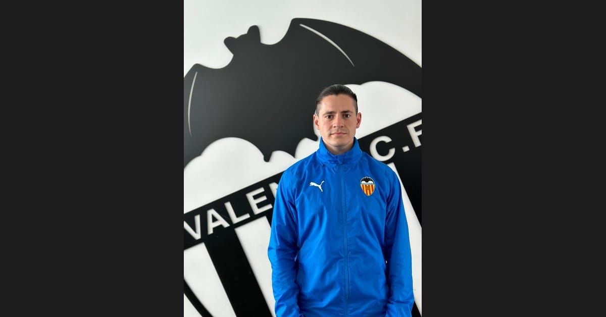 Nacho Alcaraz, Spanish coach and analyst, stands in a blue Valencia CF training jacket in front of the club's bat crest wall at the Valencia CF academy facility in Spain.