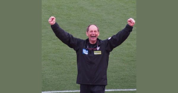 Gert Engels, German coach, in a black coaching jacket with both arms raised and laughing, celebrating a result on a Japanese football pitch.