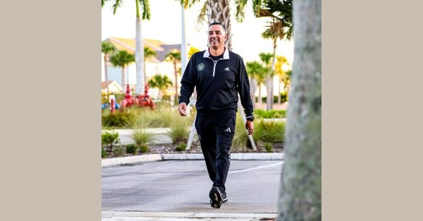 Reinerio Márquez, Chilean head coach, in a black Fort Myers City FC training top and adidas trousers, walking outdoors amid palm trees with a warm smile.