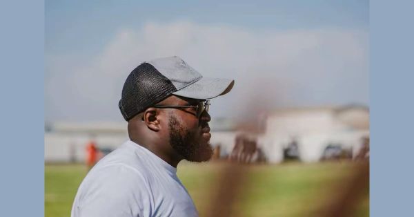 Mark Jackson, Namibian football coach, seen in profile pitchside in a cap and sunglasses, watching a match under a bright sky.