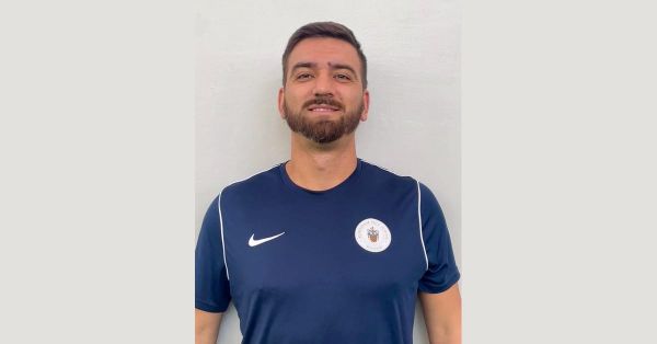 Simone Canepa, Italian football coach, in a navy Aldenham Prep School polo shirt, smiling in a head-and-shoulders portrait against a plain light wall.