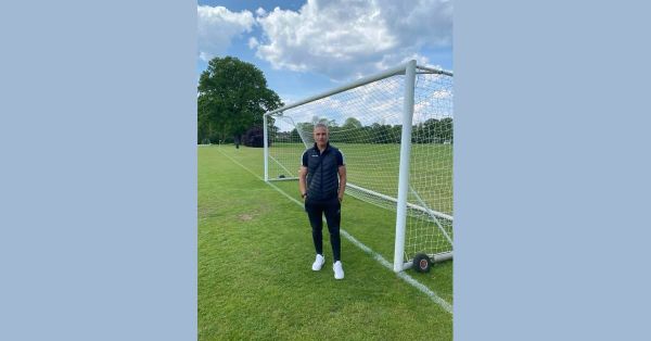 Lefteris Kalogirou, Greek football coach, standing in front of a goal on a grass pitch in a black gilet with hands in his pockets, under a partly cloudy sky.