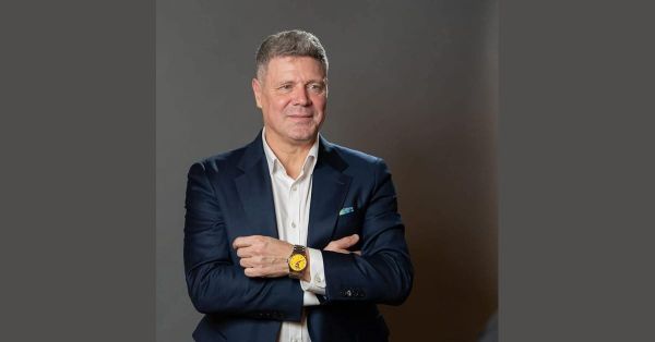 Tibor Selymes, Romanian football coach and former international, in a navy suit over a white shirt with arms crossed, in a studio portrait against a grey background.