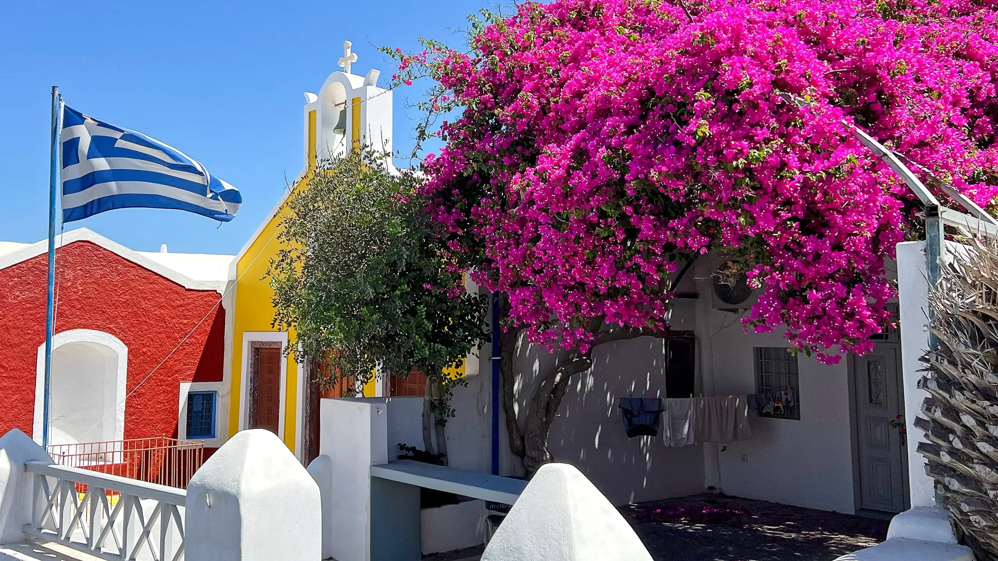 Flying greek flag, yellow church facade, with vibrant pink bougainvillea growing before them