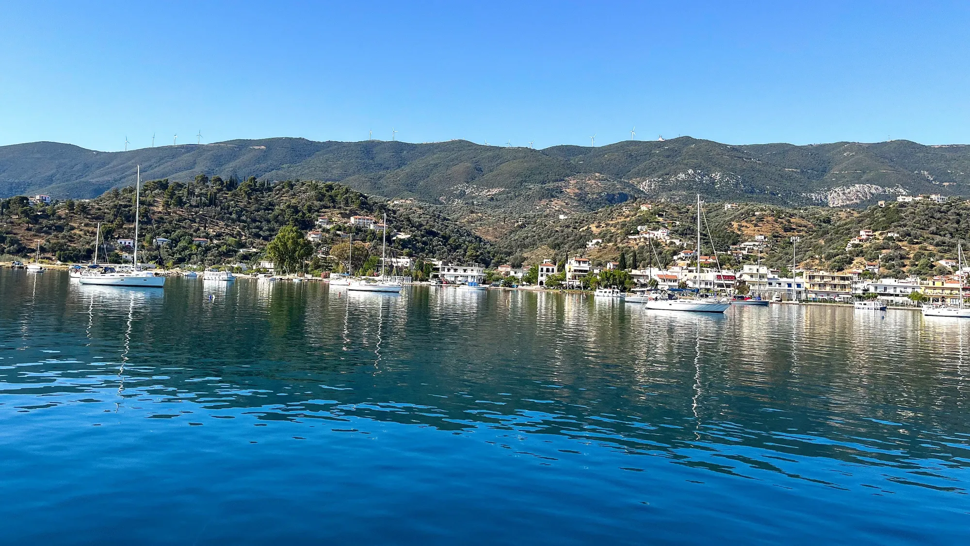 Series of boats scattered across a blue harbor set against a green hilltop