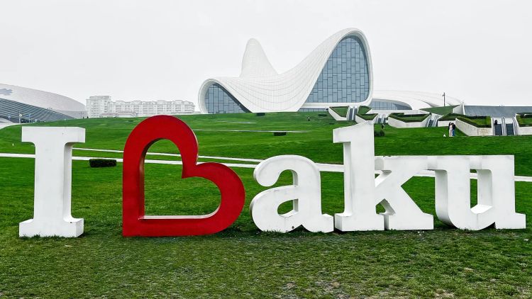 A three dimensional "I Heart Baku" sign in white located on a grassy lawn in front of a curved white building