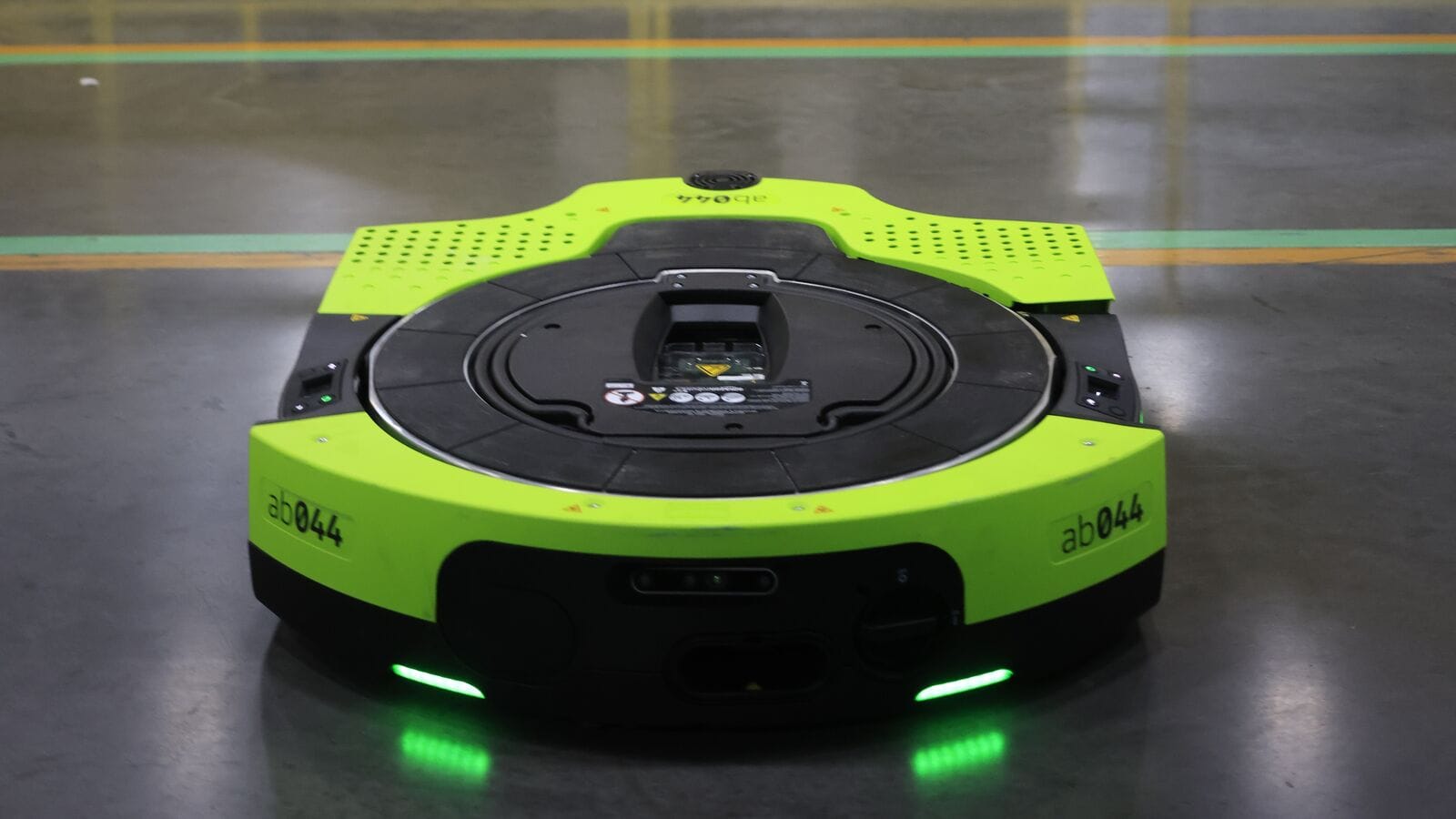 A green Amazon Proteus autonomous mobile robot on a warehouse floor with safety lights visible.