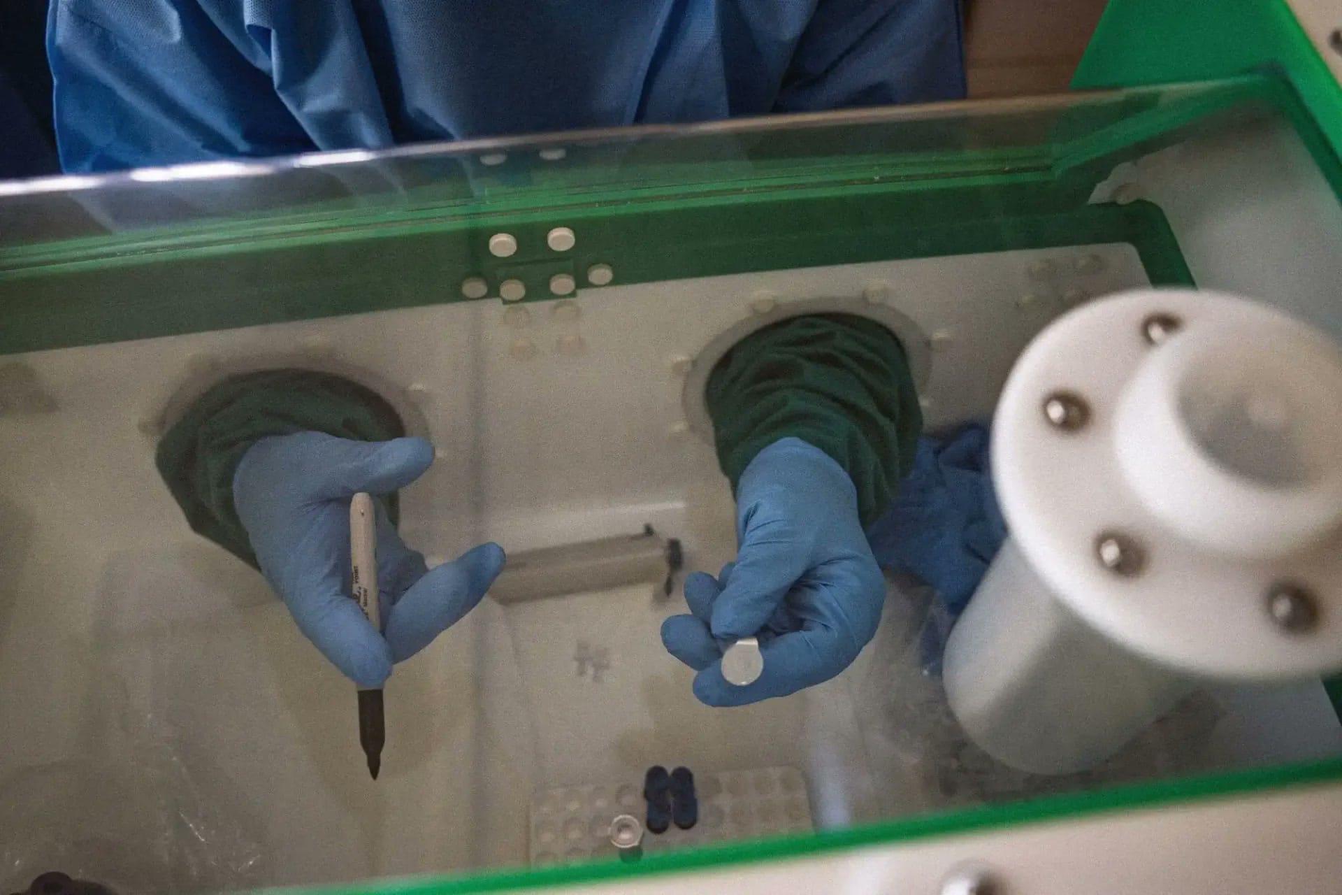 Lab worker handling samples through sealed glove ports inside a green-framed glovebox.