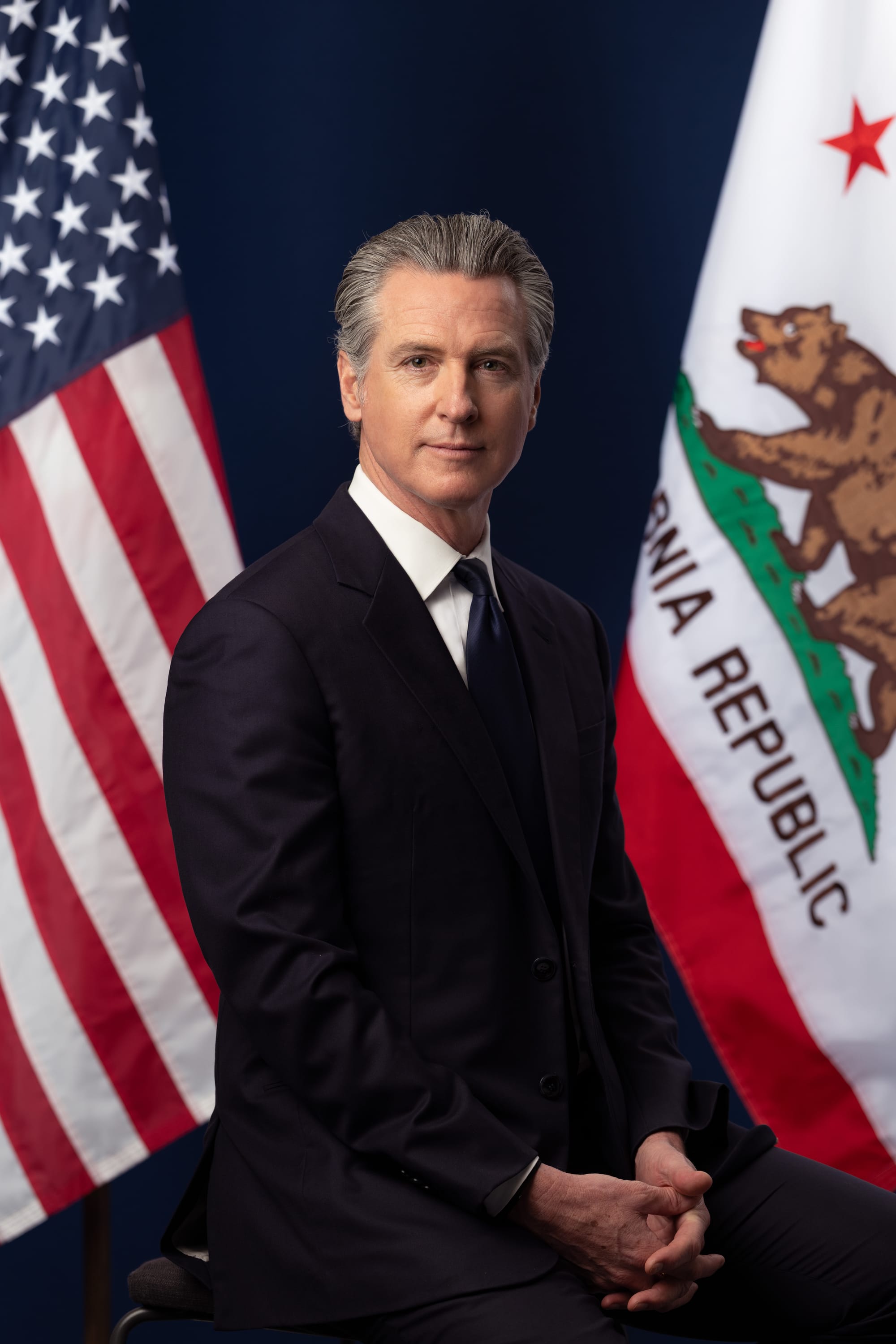 Newsom sits in a dark suit between the United States and California flags.