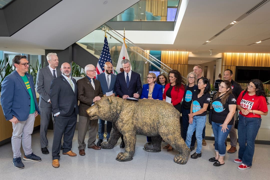 Newsom signs a document behind a golden bear statue, surrounded by a group at an indoor event.