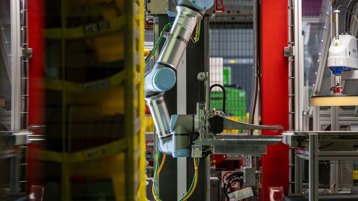 Amazon's Vulcan robotic arm and end-of-arm tool positioned beside a crowded warehouse inventory pod.