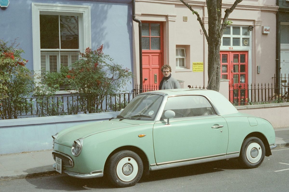 A mint-green 1990s Nissan Figaro parked in front of pastel terraced houses with red doors, shot on film.