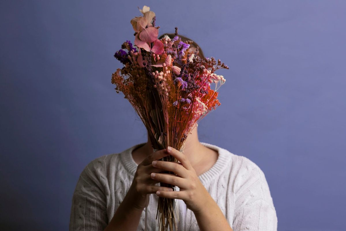 A woman in a white jumper holds a bouquet of dried pink and purple flowers in front of her face, against a lilac wall.