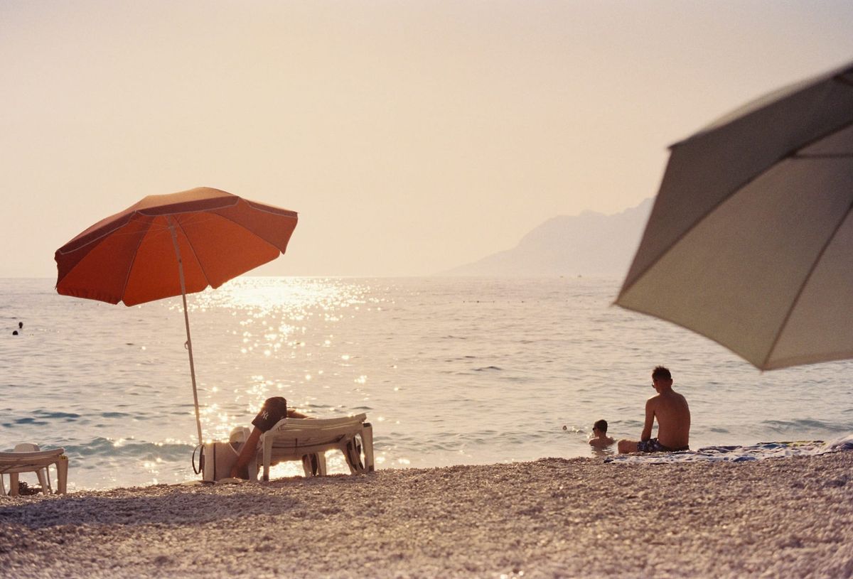 Two beach umbrellas, one orange and one white, on a pebble beach at sunset with people in the shallows, shot on film.