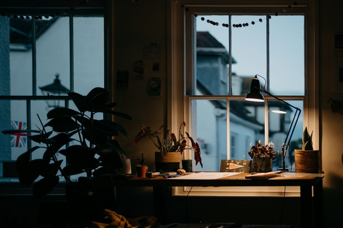 A desk lamp switched on in a dim room at dusk, with houses visible through the window behind it.