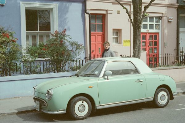 A mint-green 1990s Nissan Figaro parked in front of pastel terraced houses with red doors, shot on film.