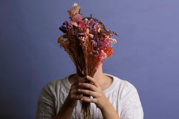 A woman in a white jumper holds a bouquet of dried pink and purple flowers in front of her face, against a lilac wall.