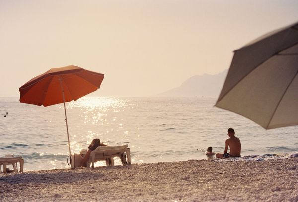 Two beach umbrellas, one orange and one white, on a pebble beach at sunset with people in the shallows, shot on film.