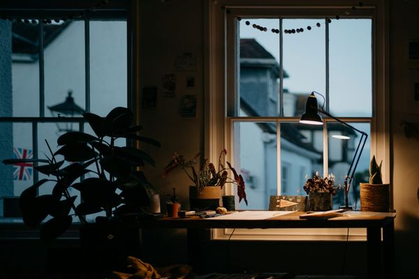 A desk lamp switched on in a dim room at dusk, with houses visible through the window behind it.