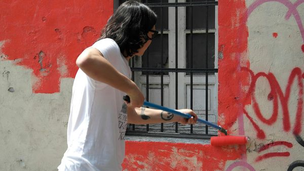 A painter on a long roller turns a pink wall white, with a blue shuttered window, a ladder and a barrier in front, shot on film.