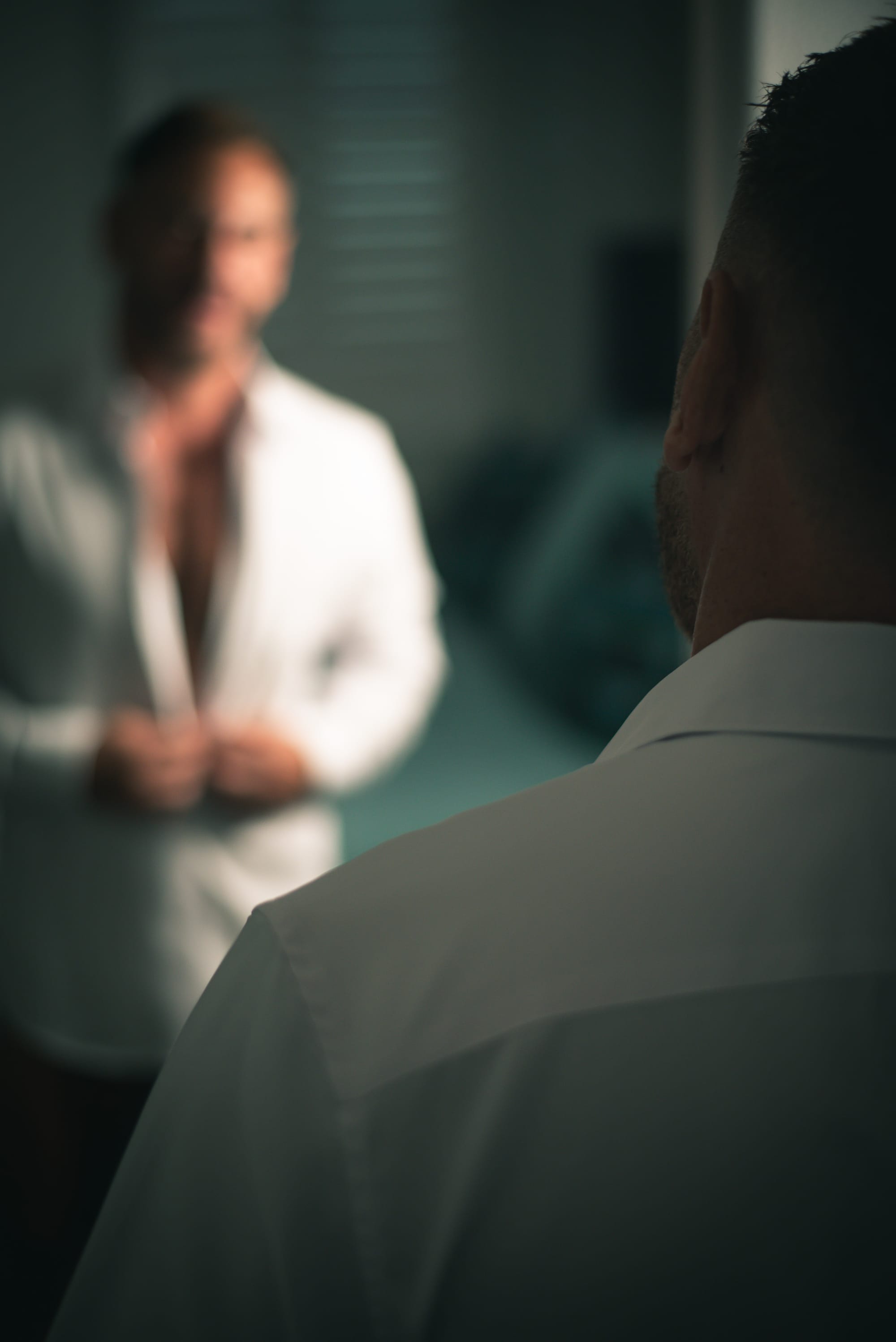 We look over David's shoulder as he unbuttons a crisp white dress shirt while looking at himself in a mirror.