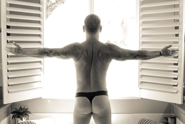 David faces a bright window with shutters thrown open. His back muscles flex as the light wraps warmly around him. He wears nothing except a black thong, its wide black waistband framing.