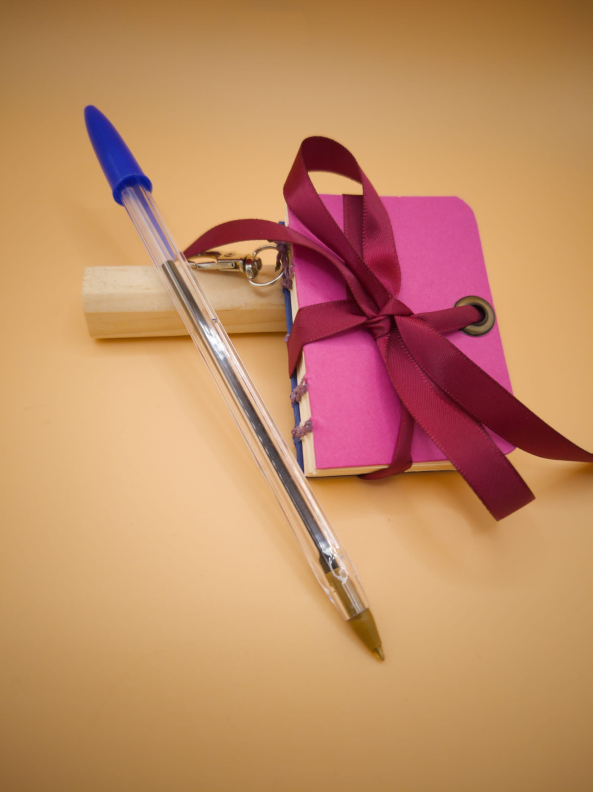 Mini carnet artisanal rose fuchsia et bleu, des coutures violettes, posé contre un bout de bois, sur un fond beige orangé, avec un ruban bordeaux noué dessus, un petit mousqueton accroché, et un stylo bille appuyé contre lui (le stylo est plus grand que le carnet)