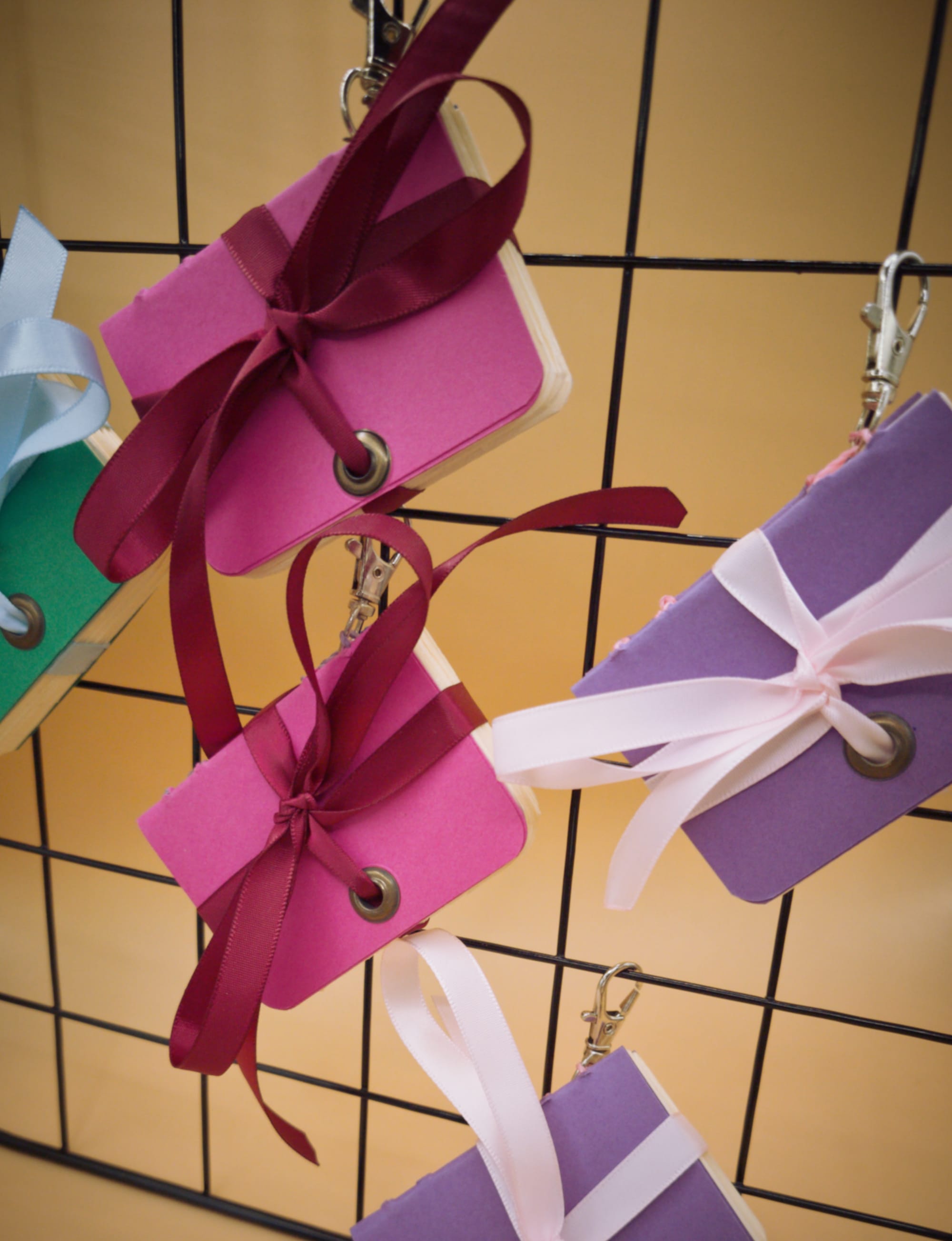 Gros plan sur des mini carnets artisanaux suspendus à une grille noire deux en rose fuchsia avec ruban bordeaux, deux en violet avec ruban rose cadrés serrés, et un vert sapin à ruban bleu pâle