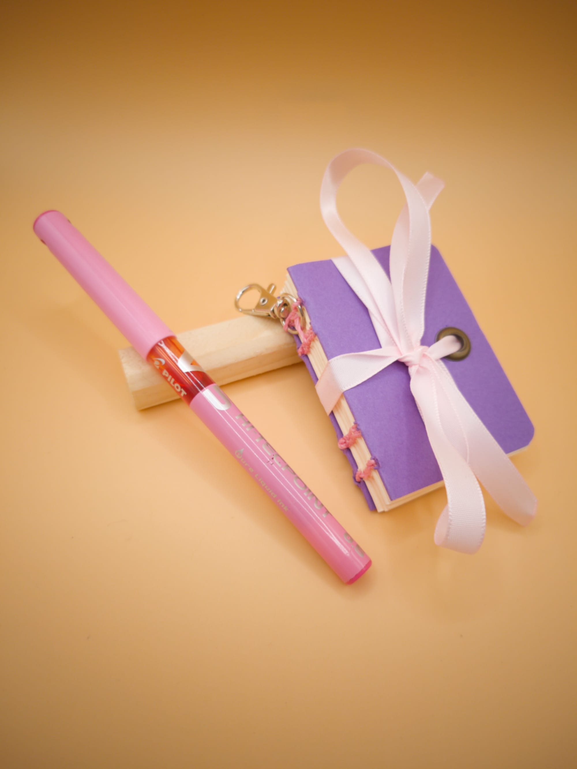 Mini carnet artisanal lilas à coutures rose pâle, posé contre un bout de bois, sur un fond beige orangé, avec un ruban rose pâle noué dessus, un petit mousqueton accroché, et un stylo rose appuyé contre lui (le stylo est plus grand que le carnet)