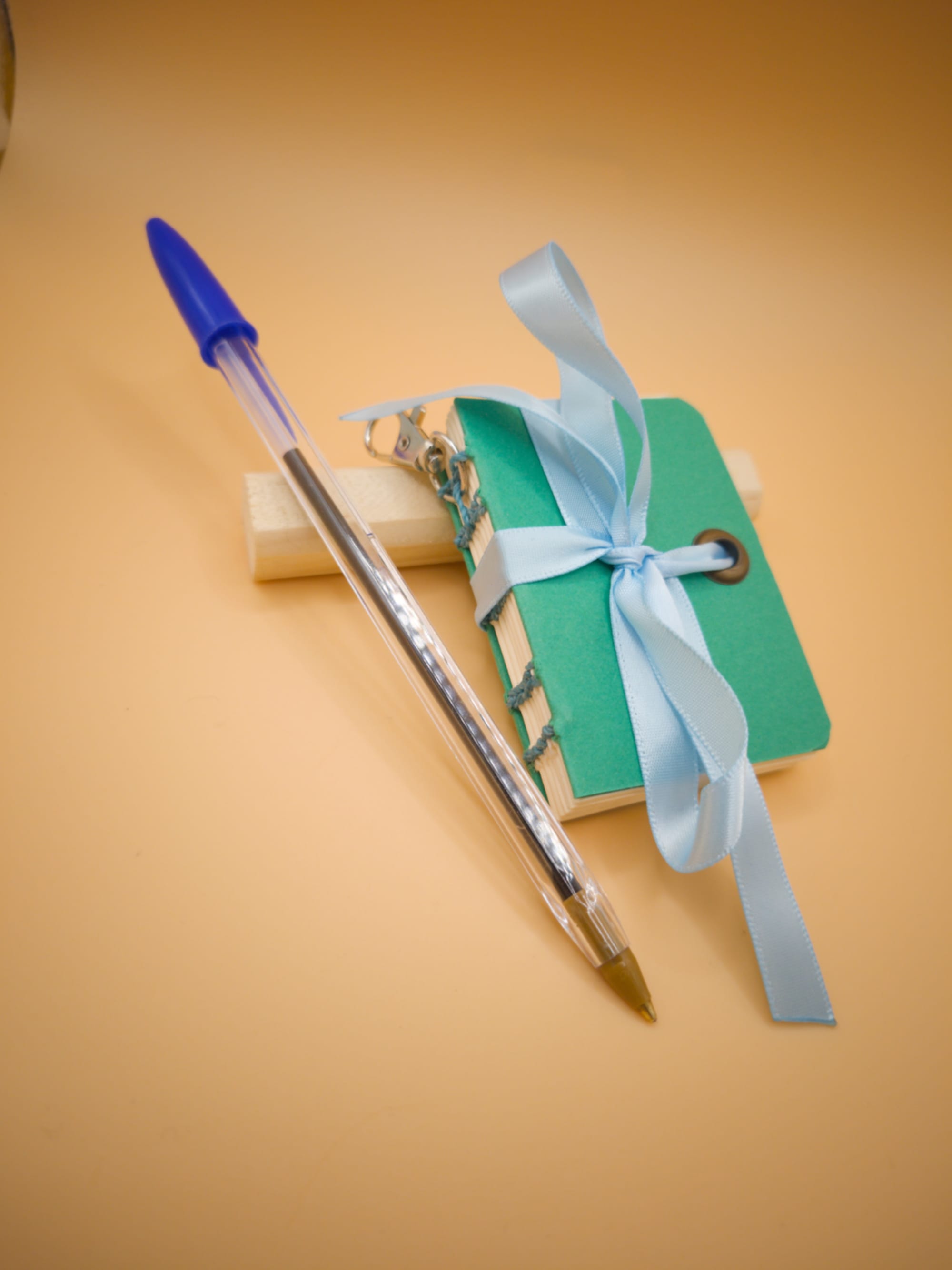 Mini carnet artisanal vert sapin à coutures bleues clair, posé contre un bout de bois, sur un fond beige orangé, avec un ruban bleu pâle noué dessus, un petit mousqueton accroché, et un stylo bille appuyé contre lui (le stylo est plus grand que le carnet)