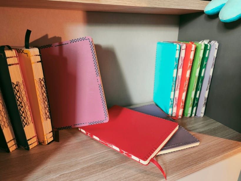 Des carnets de toutes tailles et de toutes épaisseurs, reliés à la main avec des couleurs variées et des motifs de fils apparents différents, posés sur un meuble en bois