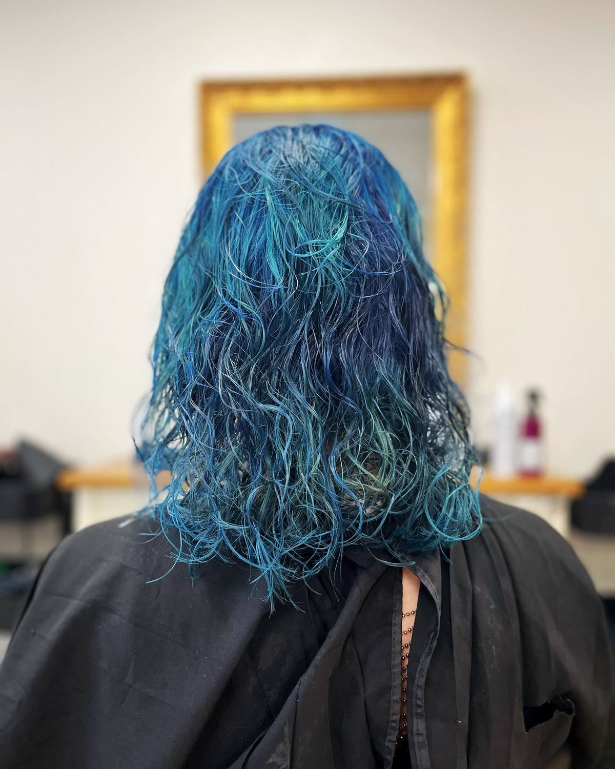 Mes cheveux vus de dos dans un salon de coiffure/coloration, ils sont bleus avec des reflets plus sombres, et des ondulations fines et des légères boucles.