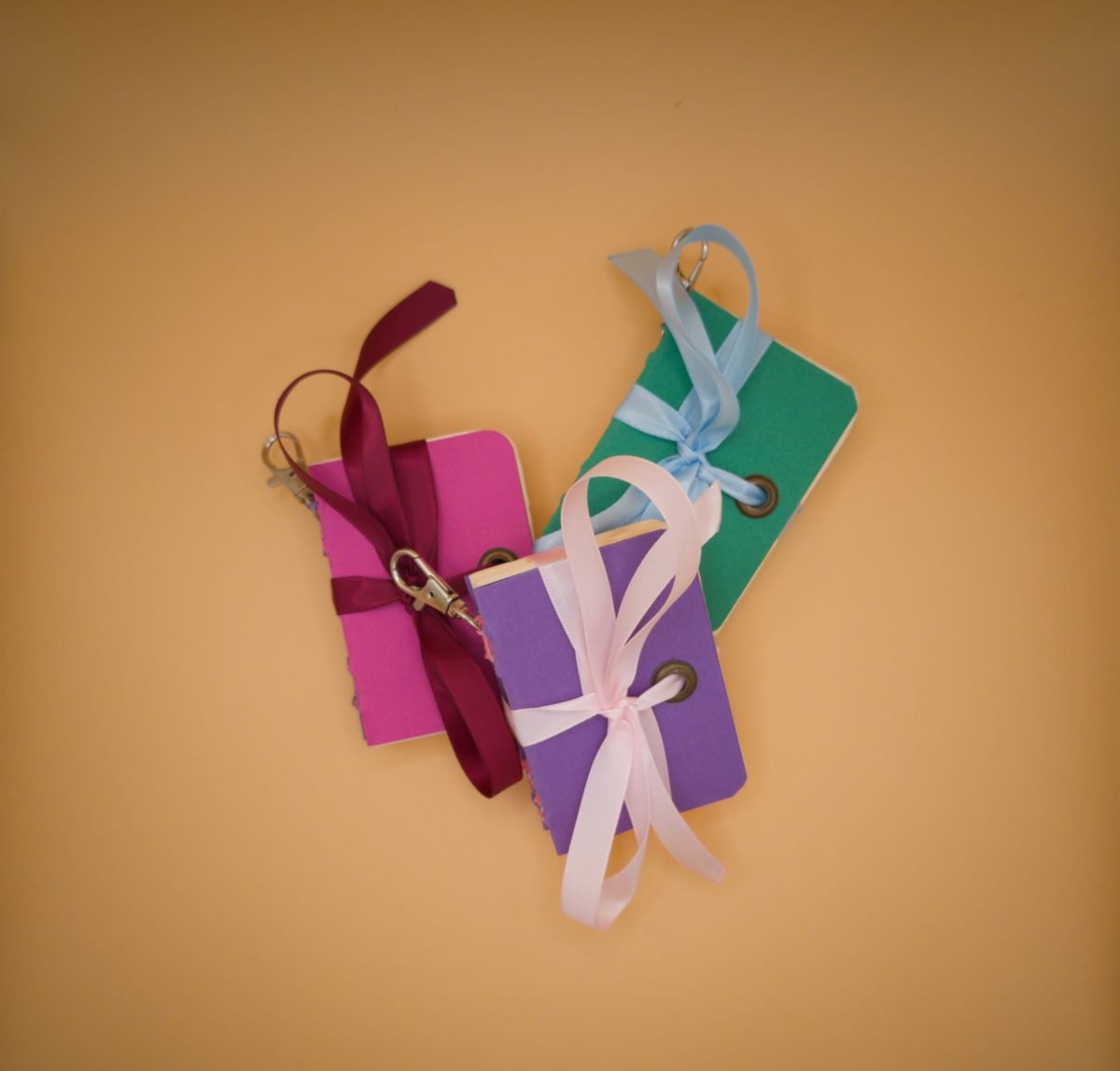 Trois mini carnets artisanaux éparpillés sur fond beige orangé : rose fuchsia avec ruban bordeaux, violet avec ruban rose pâle et œillet, vert sapin avec ruban bleu ciel