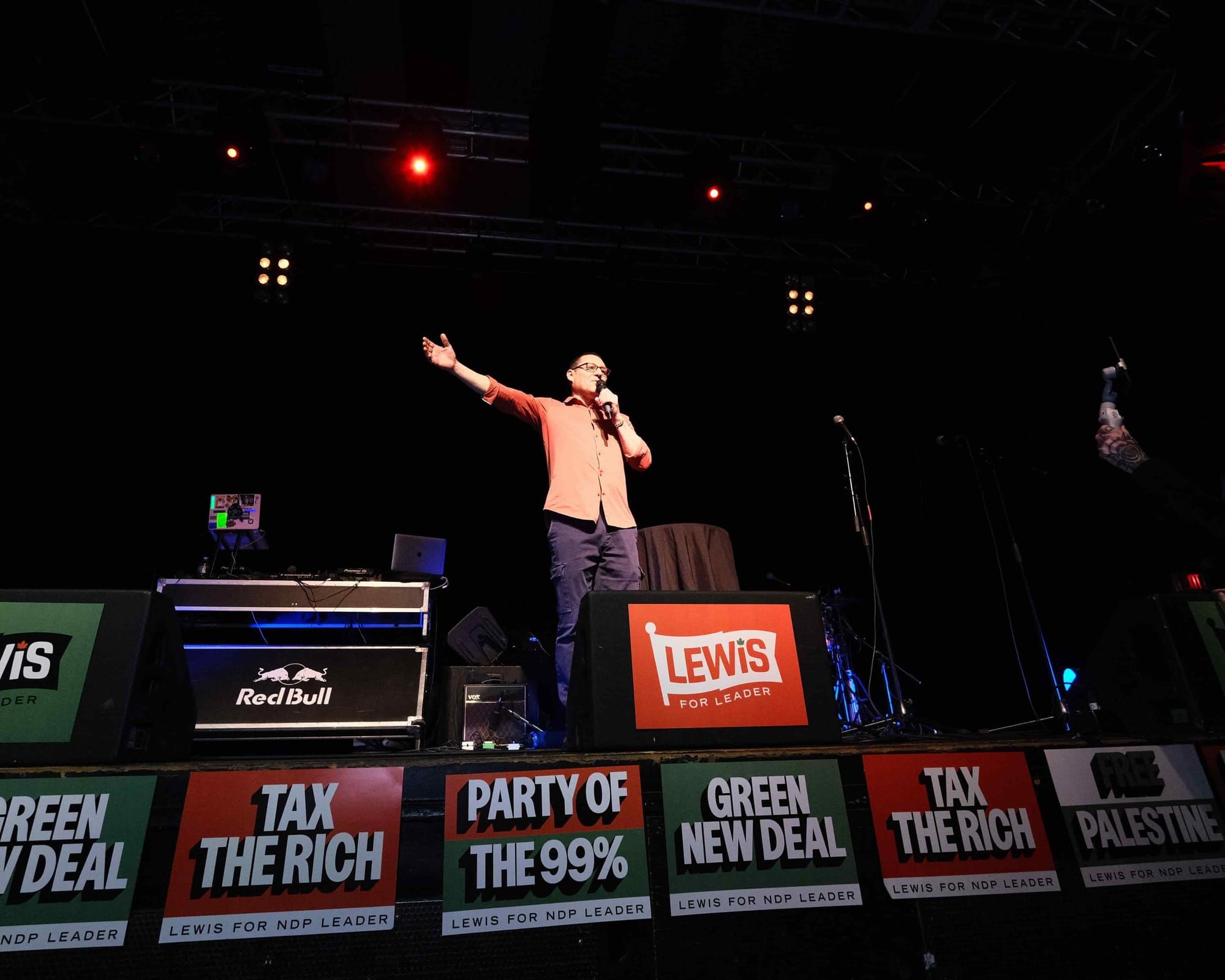 A photo of Avi Lewis giving a speech on a stage with campaign signs in front of him