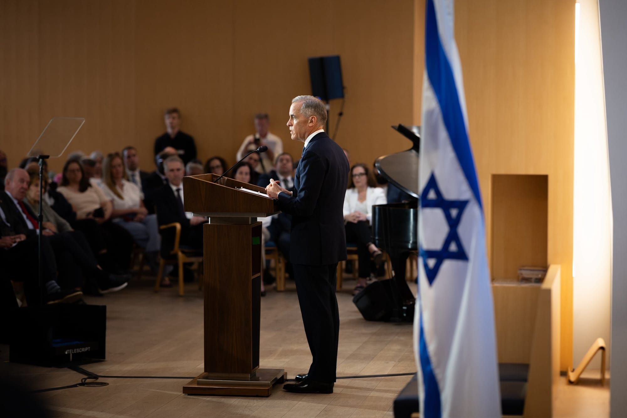 Mark Carney giving a speech in front of an Israeli flag