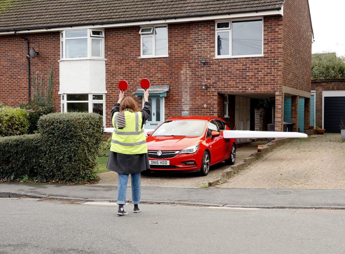An aircraft marshall guides a car with wings taxi on the runway in front of a suburban house