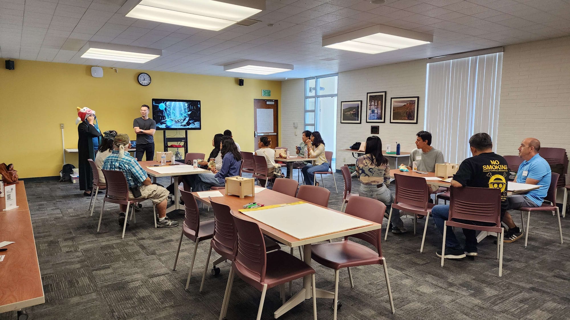 Meeting room with 4 large tables with 4 people seated at each table. At the front of the room stands a wizard wearing a fish hat and another person standing in front of a large TV screen.